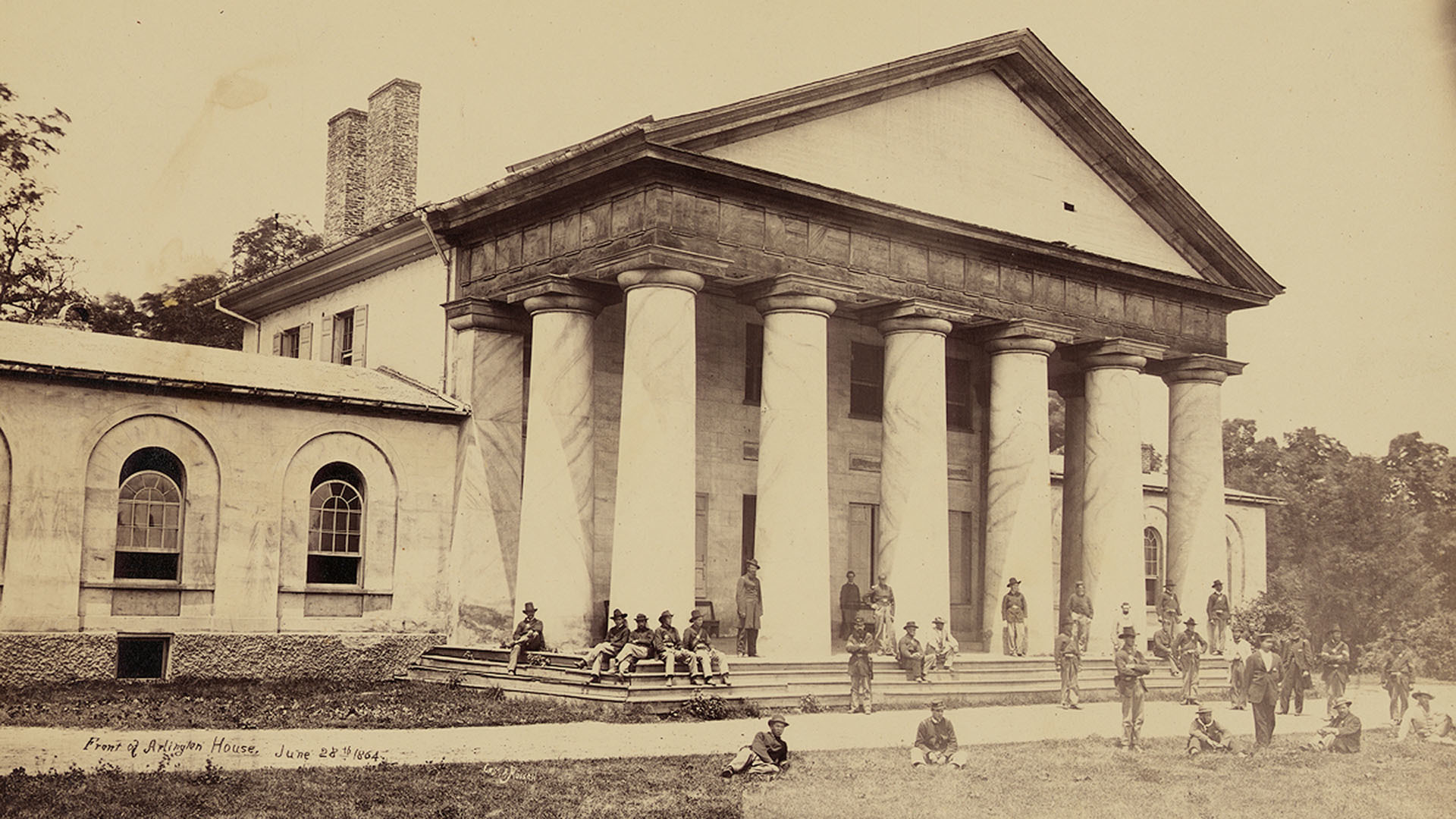 A sepia-toned photo of Union soldiers gathered outside Robert E. Lee's large home, which has grand columns outside it, in Arlington, Virginia, circa 1864.