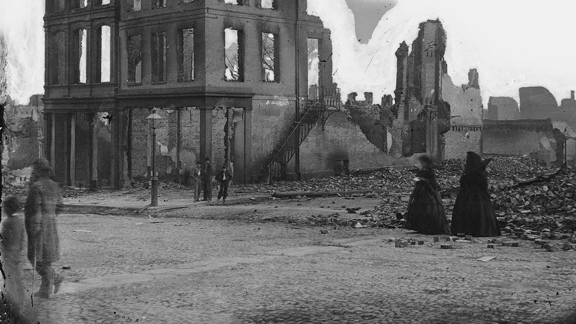 A black-and-white photo showing men and women walking past a ruined building in Richmond, Virginia, circa 1865.