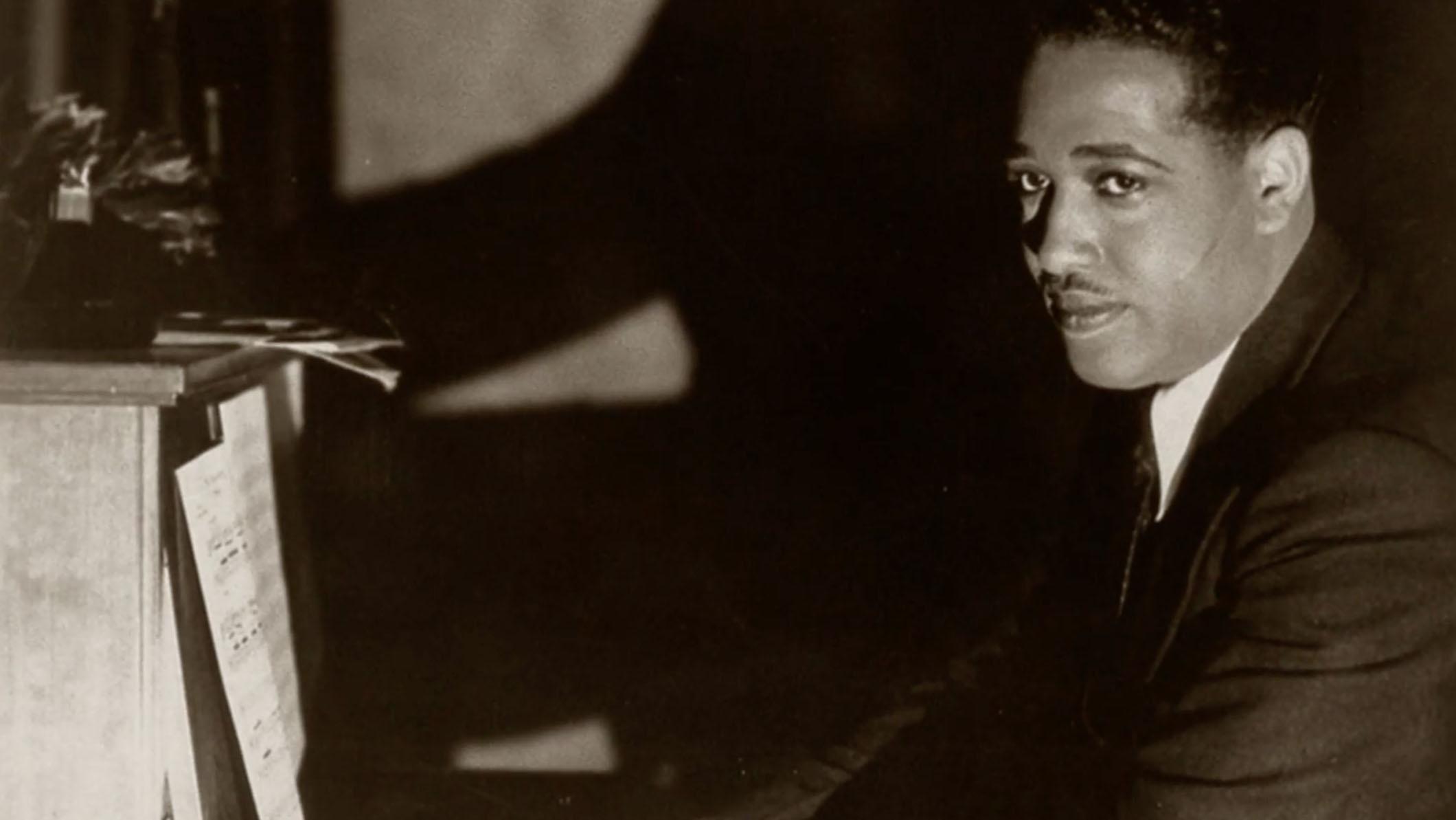 A black-and-white photo of a man posing for a photo, seated at a piano.