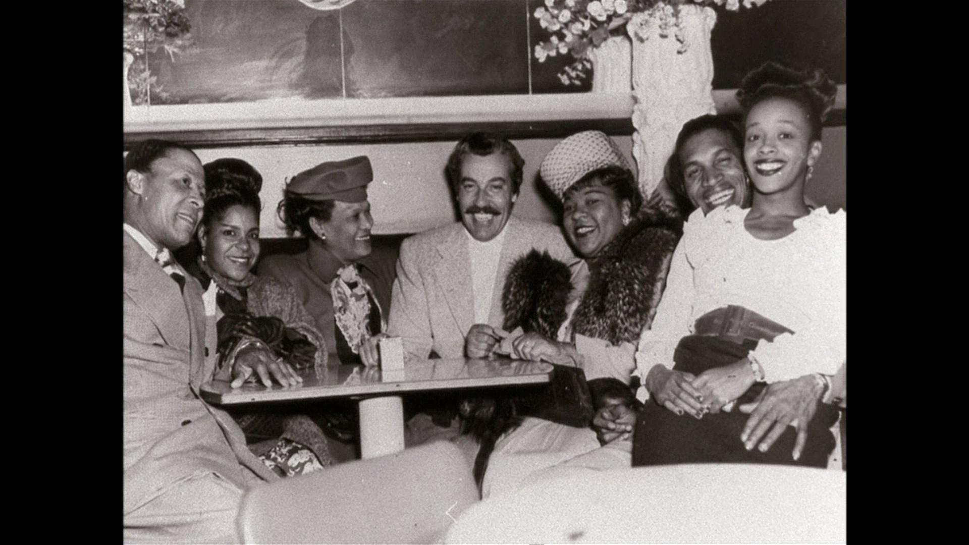 A black-and-white photo of a group of people seated around a table, they are all smiling.