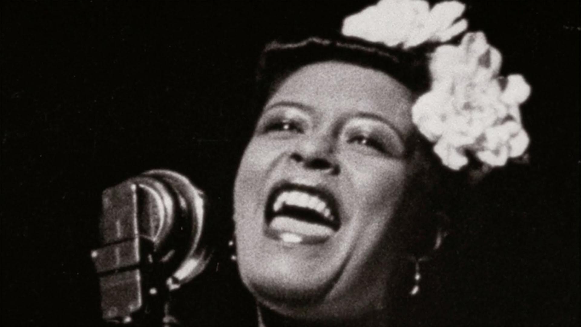 A black-and-white photo of a woman with folowers in her hair singing into a microphone.