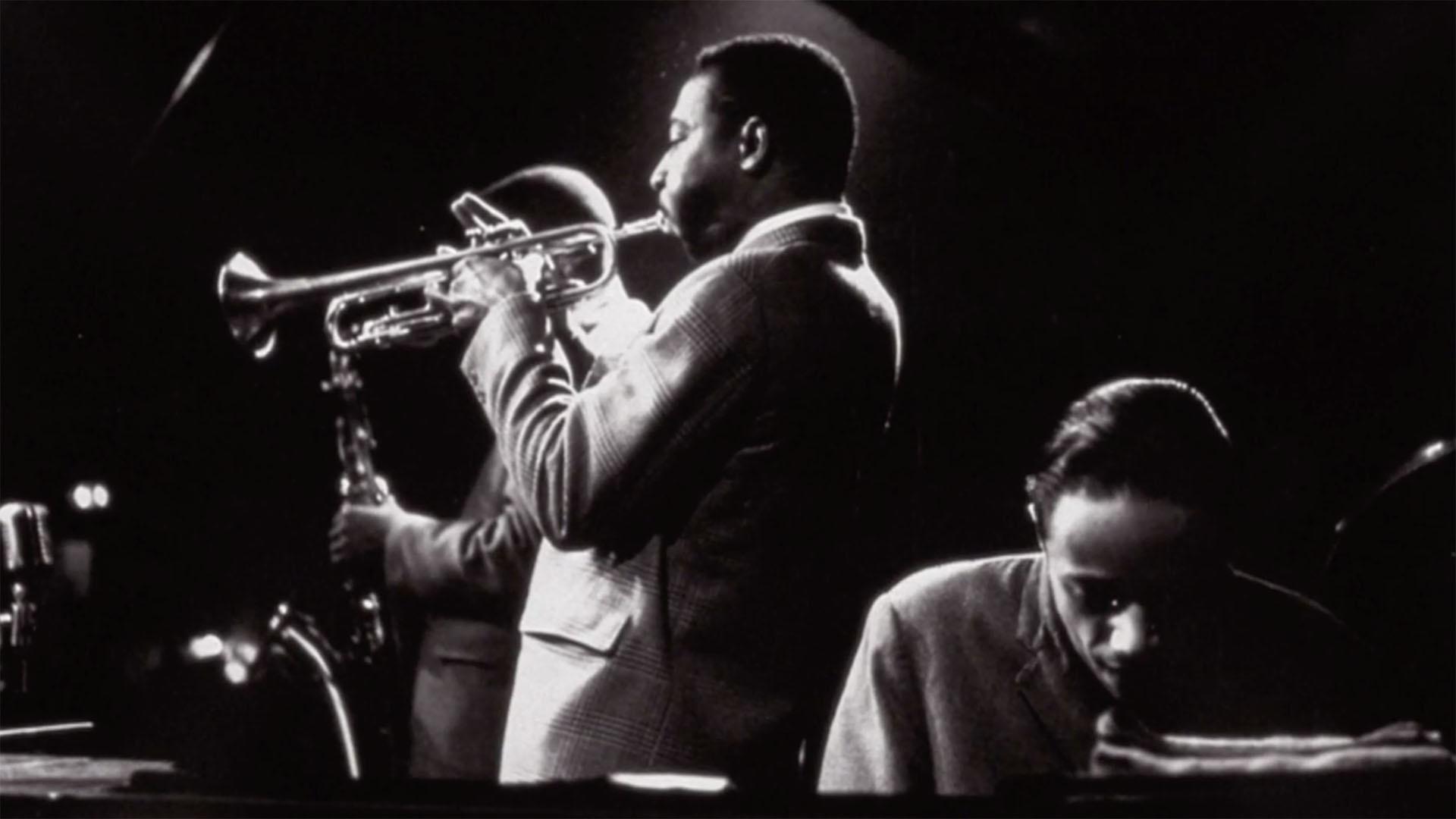 A black-and-white photo of a man facing away from the camera, playing a trumpet.