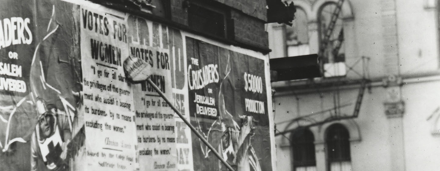 A black-and-white photo that has been split into three pieces horizontally. This is the first of three. It shows two women in early twentieth century garb plastering posters on a wall which call for women's right to vote.
