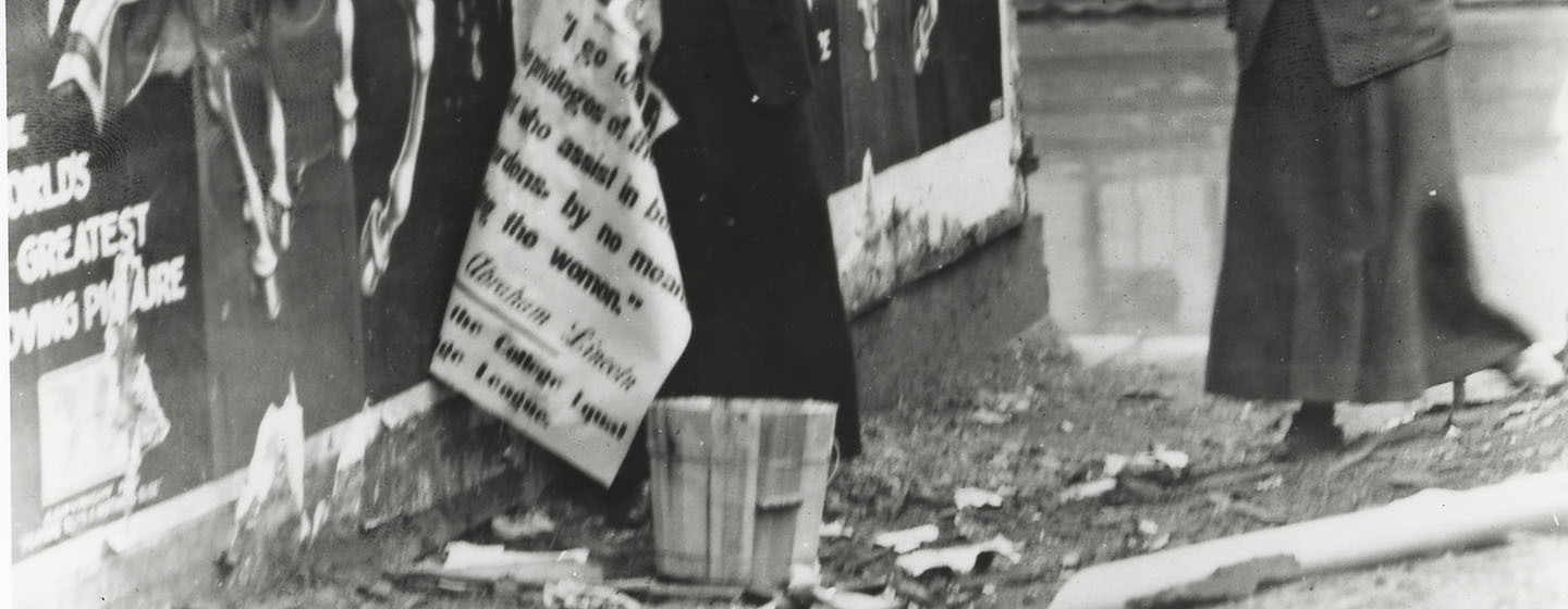 A black-and-white photo that has been split into three pieces horizontally. This is the last of three. It shows two women in early twentieth century garb plastering posters on a wall which call for women's right to vote.