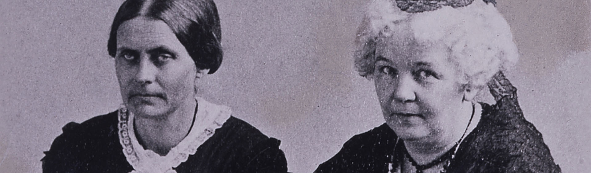 A lilac-hued photo of Elizabeth Cady Stanton sitting alongside Susan B. Anthony. They both have serious expressions on their faces.