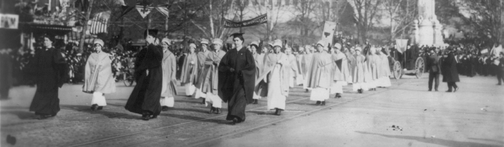 A black-and-white image with has been split into two pieces horiontally. This is the second piece. The image depicts a procession of female nurses in 1913, in Washington D.C., as they hold banners aloft which proclaim support of the suffragettes.