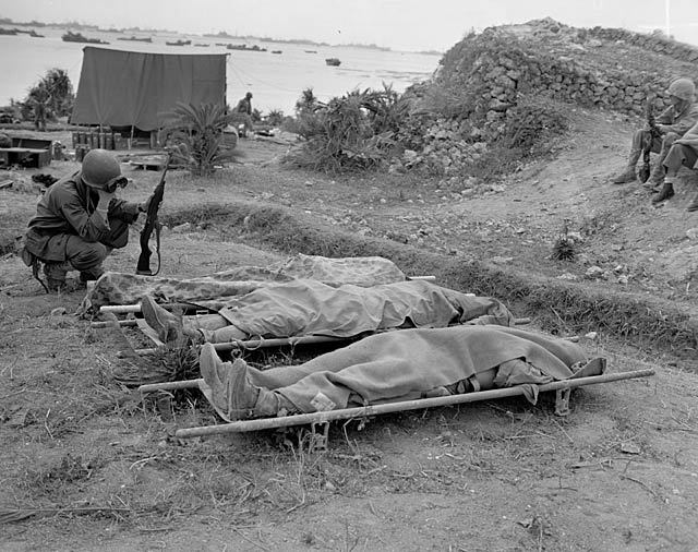 A lone Marine crouches solemnly above a row of three covered American bodies on Okinawa. April 4, 1945.