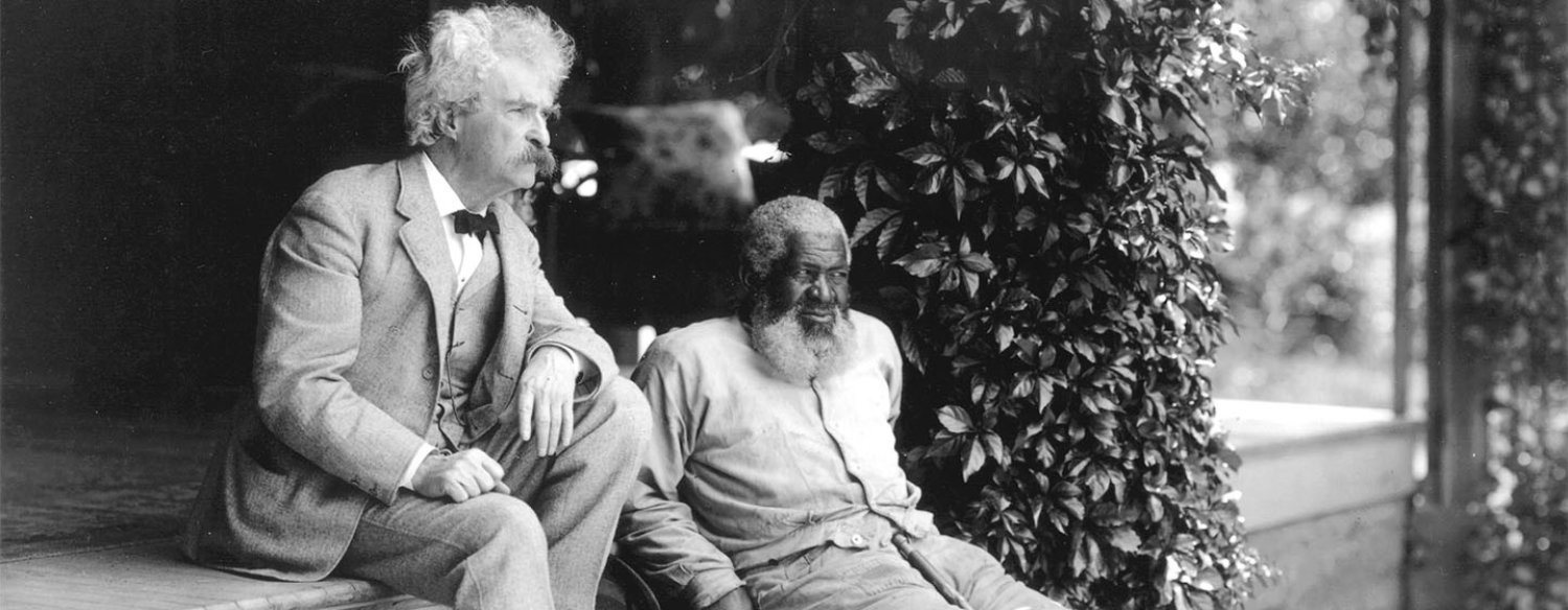 A black-and-white photo of Mark Twain wearing a pale suit and bow tie, his hair and beard white in color, sitting on some outdoor steps next to John Lewis, an elderly African American man with a large white beard.