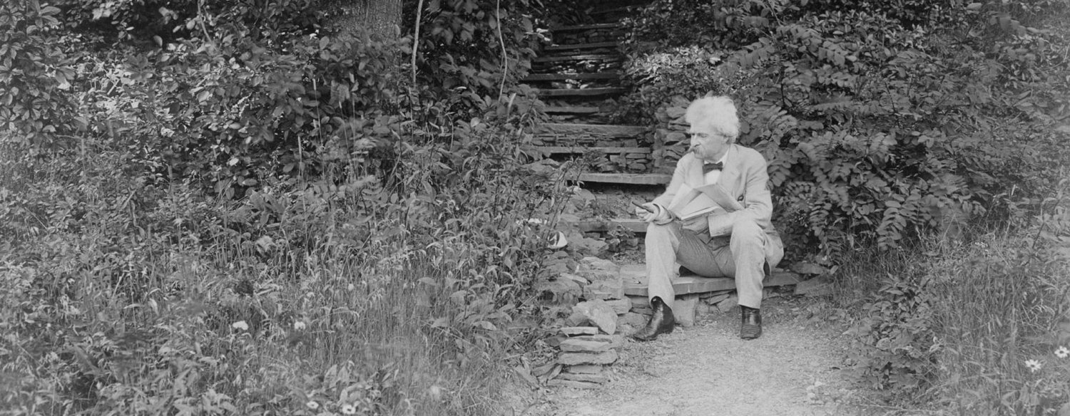 A black-and-white photo of Mark Twain sitting on outdoor steps along a secluded garden path, surrounded by greenery. He is looking away from the camera.