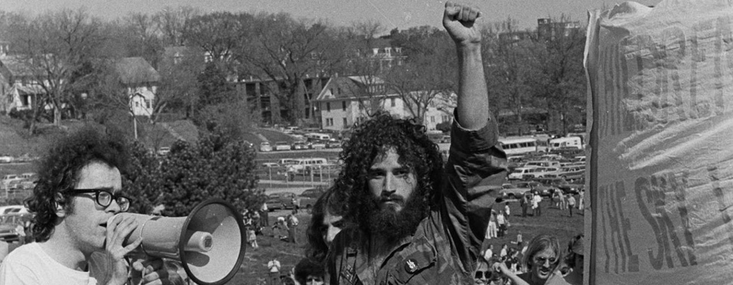 A black-and-white photo of American war protestors during the Vietnam era. One man wearing a military-style camouflage jacket holds his fist aloft in the air while a man next to him is speaking into a bullhorn.