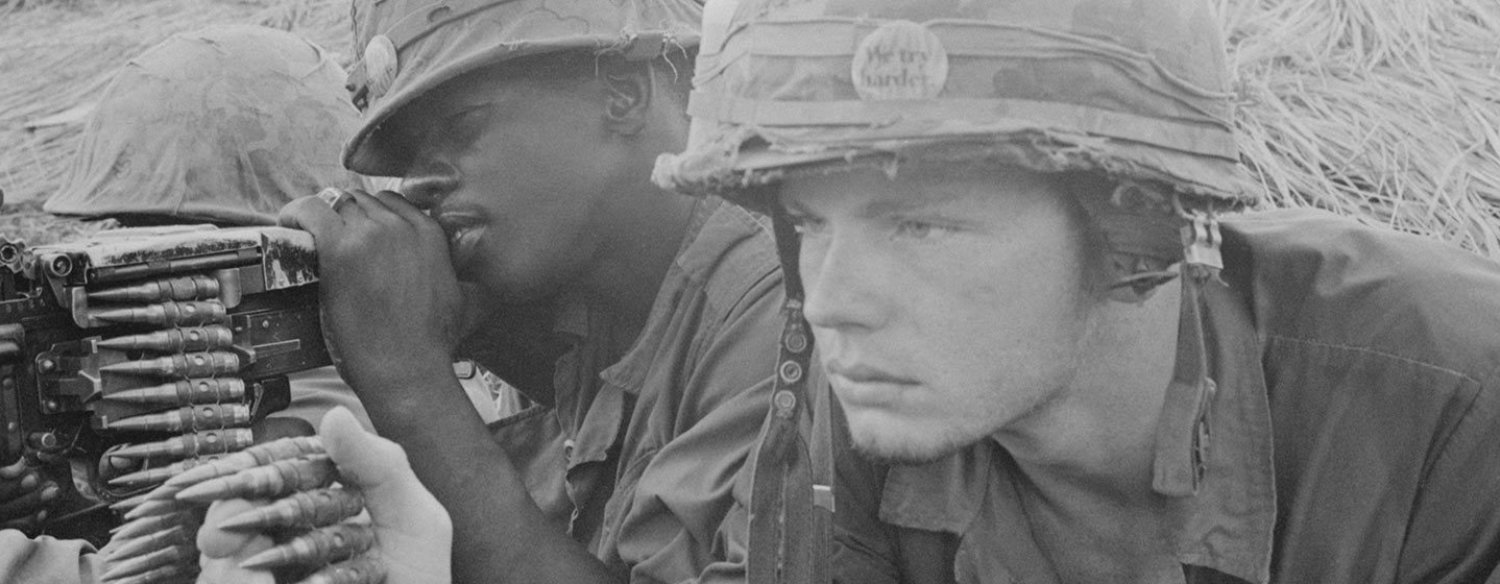 A black-and-white photo of three American soldiers, laying prone. One man is looking down the the sight of a machine gun. The other man wears a camouflage helmet with a pin on it that says "we try harder." Both men have terse expressions.