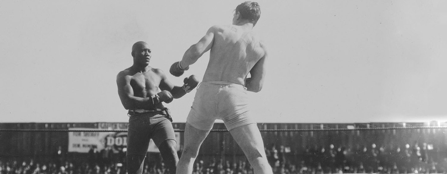 A black-and-white photo of Jack Johnson in a wrestling ring, fighting an opponent. Johnson's opponent has his back to the camera and Johnson is squaring up against him, facing the camera. There are many people in the crowd.