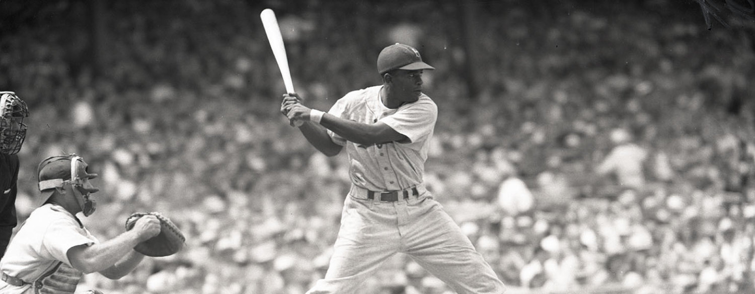 A black-and-white photo of Jackie Robinson batting during a baseball game. He is mid-swing.