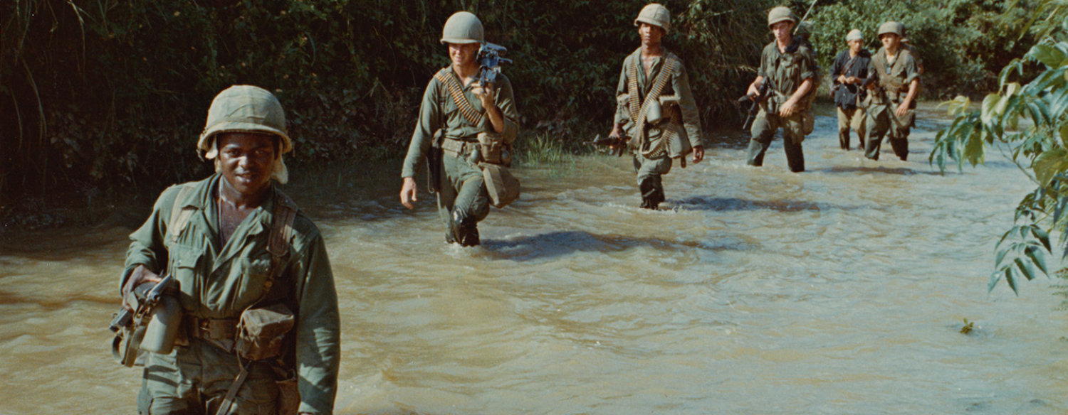 A color photo of Vietnam-era American military personnel trudging through thigh-deep water, in a single-file line, in the Vietnamese jungle.
