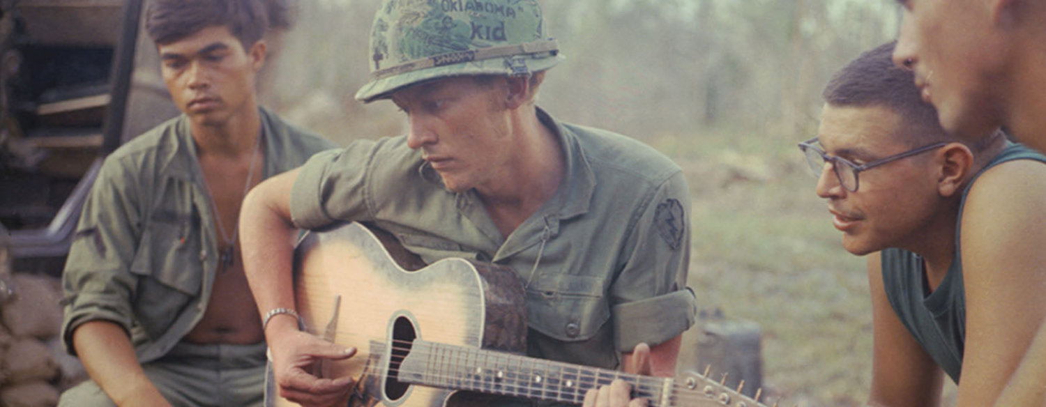 A color photo of an American serviceman playing an acoustic guitar as several other military members gather around to listen. The man playing the guitar wears a camouflage helmet with the words "Oklahoma Kid" scrawled on it.