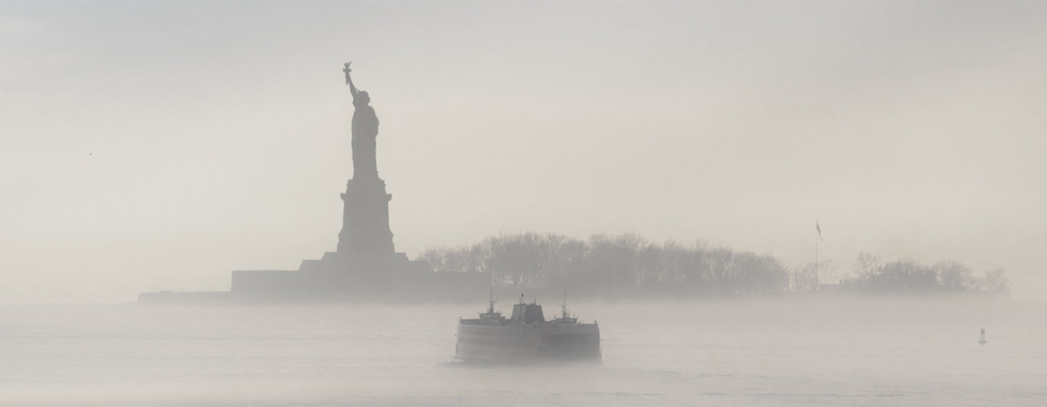 A photo of The Statue of Liberty, which stands in the background as a boat traverses fog in the foreground.