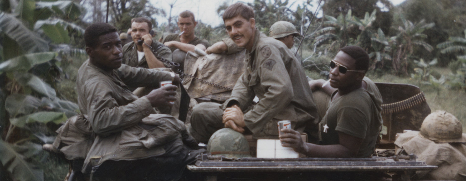 A color photo of several American servicemen from the Vietnam era sitting in a jeep in a dense jungle, looking back at the camera.