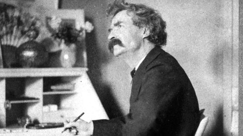 A black-and-white photo of Mark Twain, seated at his desk with a pen in hand. He is looking up from the desk, lost in thought. | Selected Writings
