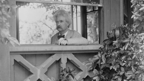 A black-and-white photo of Mark Twain, inside a house, looking out of a window which is partly framed by ivy and flowers. | About Twain