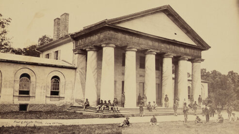 A sepia-toned photo of Union soldiers gathered outside Robert E. Lee's large home, which has grand columns outside it, in Arlington, Virginia, circa 1864. | Episode 7 | Most Hallowed Ground (1864)