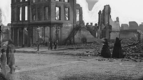 A black-and-white photo showing men and women walking past a ruined building in Richmond, Virginia, circa 1865. | Episode 8 | War Is All Hell (1865)