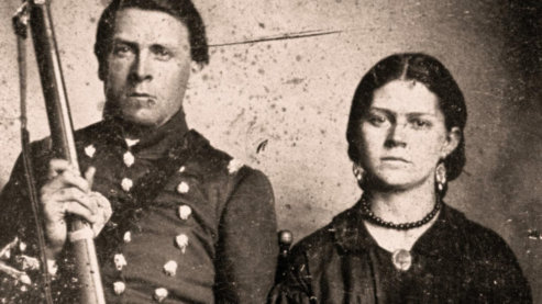 A sepia-toned photo of a man in soldier's garb posing for the photo next to a woman in a black dress, wearing a locket around her neck. The man holds a rifle upright. They both have serious expressions. | Video