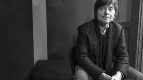 A black-and-white photo of Ken Burns seated, looking at the camera. | Q&A with Ken Burns