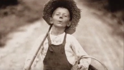 A sepia-toned photo of a young boy walking down a rural dirt road. He is wearing overalls and a wide-brimmed straw hat. He carries a fishing pole over his shoulder and a woven basket under one arm. | Video