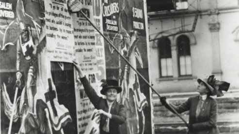 A black-and-white photo that shows two women in early twentieth century garb plastering posters on a wall which call for women's right to vote. | Women's Suffrage