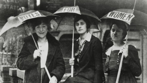 A black-and-white photo of three women looking at the camera as they carry open umbrellas. Each umbrella has one word on it which spells out "Votes For Women." | Women Remember Suffrage