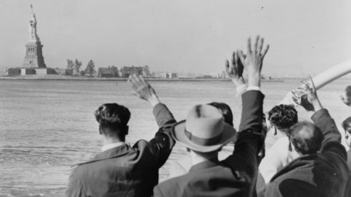 A black-and-white photo of a group of men waving goodbye to the Statue of Liberty in the distance, as their boat sails away. | Timeline