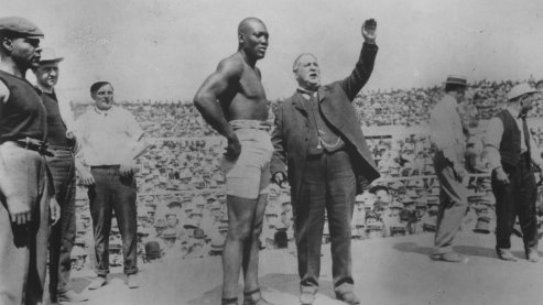 A black-and-white photo of Jack Johnson in a boxing ring, standing next to an announcer, as other men also stand nearby. The announcer has his hand raised above his head. | Timeline