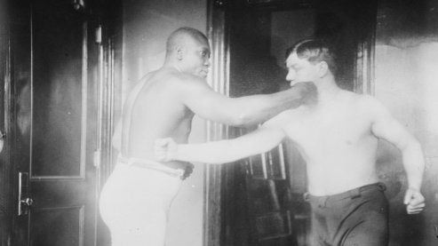 A black-and-white photo of Jack Johnson posing with another boxer. They are both pantomiming punching one another. The other boxer's fist is connecting with Johnson's chest while Johnson is connecting with his opponent's jaw. | Boxing Glossary