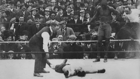 A black-and-white photo of Jack Johnson standing in a boxing ring, over a prone, defeated opponent. The umpire is counting the opponent out and there is a large crowd in the background. | About Jack Johnson
