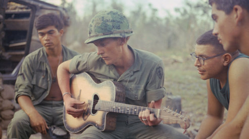 A color photo of an American serviceman playing an acoustic guitar as several other military members gather around to listen. The man playing the guitar wears a camouflage helmet with the words "Oklahoma Kid" scrawled on it. | Music