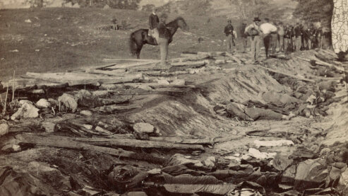 A sepia-toned photo shows men looking into a ditch on the side of a rural road, where many bodies are piled. | Civil War Facts