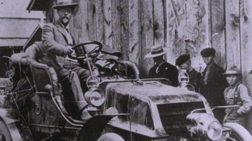 A lilac-hued photo of a man sitting atop an old-fashioned, open-air vehicle. He is dressed in formal attire. There are a line of people standing nearby wearing more simple clothing. | Ken Burns on Horatio's Drive
