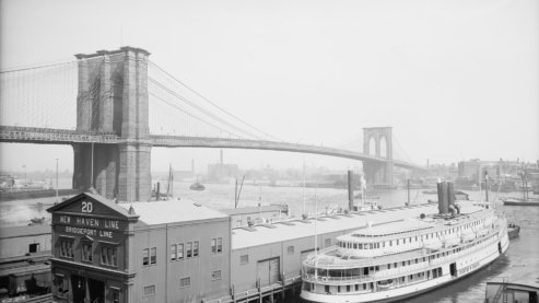 A black-and-white photo taken in the early 20th century, depicting the Brooklyn Bridge with a large ferry in the foreground. | Timeline