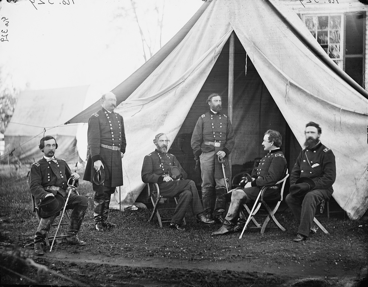 A black-and-white photo shows men in the military garb of generals, standing and sitting outside a large tent.